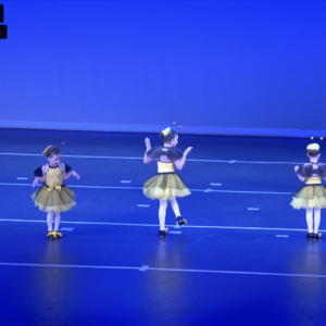 Two children in bee-themed costumes perform a dance routine on stage under blue lighting as part of the Recital Package for Two Students, moving with raised or extended arms and facing different directions.